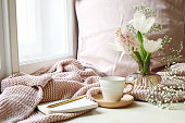 Cozy Easter, spring still life scene. Cup of coffee, opened notebook, pink knitted plaid on windowsill. Vintage feminine styled photo. Floral composition with tulips, hyacinth and Gypsophila flowers.