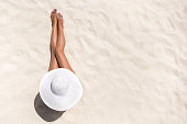 Summer holiday fashion concept - tanning woman wearing sun hat at the beach on a white sand shot from above