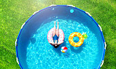 Aerial. Girl resting in a metal frame pool with inflatable toys. Summer leisure and fun concept. Frame pool stand on a green grass lawn. Top view from drone.