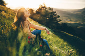 Young woman traveler with long loose curly hair sits on green grass meadow with flowers and types on smartphone against hilly landscape under sunlight