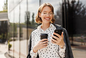 Positive business woman with take away coffee using mobile phone while standing on sunny street