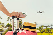 Woman with pink suitcase and passport with boarding pass standing on passengers ladder of airplane opposite sea with palm trees. Tourism concept