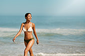 Fit, beautiful and toned bikini model on beach enjoying fun and relaxing summer holiday by ocean and sea while wearing a swimsuit. Smiling, happy and walking woman in swimwear on a tropical vacation