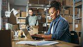 Diverse Male and Female Warehouse Inventory Managers Talking, Using Laptop Computer and Checking Retail Stock. Rows of Shelves Full of Cardboard Box Packages in the Background.