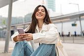 Waiting for the bus and for love. Bright cheerful young woman of Turkish mixed race in white casual clothes with a charming smile holding a phone in the street at public transportation stop