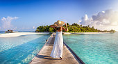 Back view of a woman in white dress walking down a pier towards a tropical island