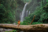 Woman sitting on huge log on the background of tropical waterfall while hiking